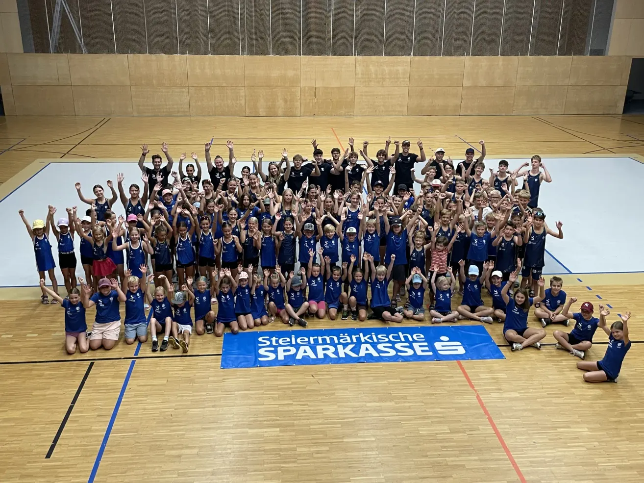 Eine Gruppe von Kindern und Erwachsenen in blauen Uniformen posiert für ein Foto in einer Turnhalle mit einem Banner, das 'Steiermarkische Sparkasse' steht.
