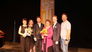 Fünf Personen stehen auf einer Bühne und lächeln für ein Foto. Eine Frau in einem rosa Oberteil hält Blumen. Hinter ihnen ist ein Banner mit dem Text 'BACH Kultur sommer'.