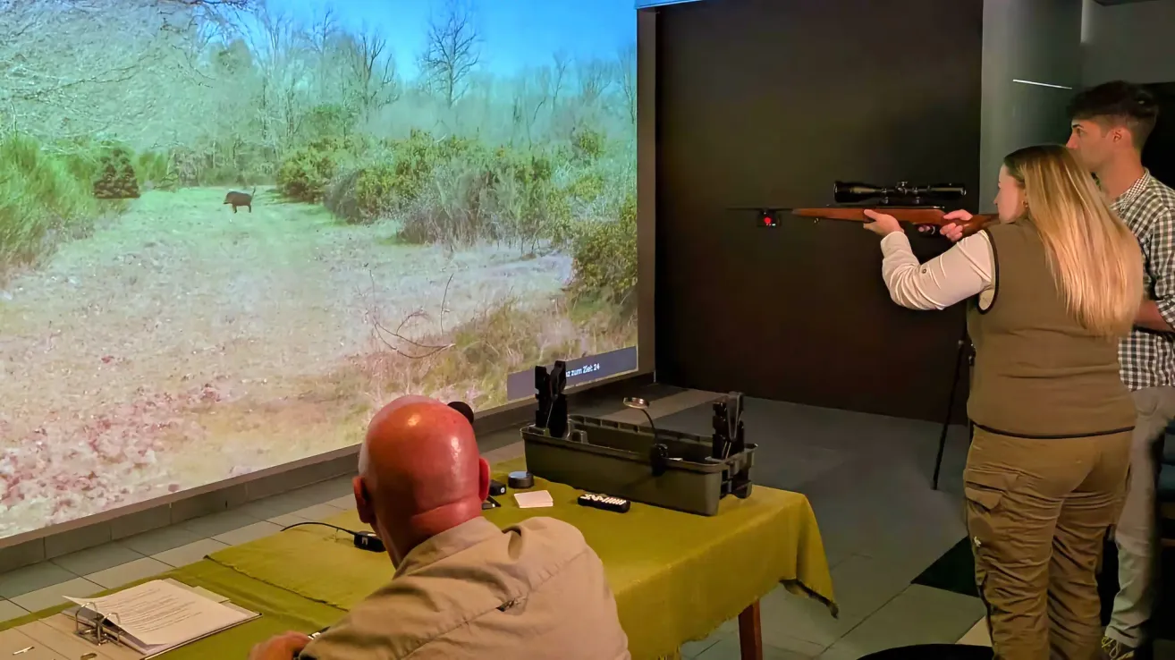 Ein Mann sitzt vor einem Bildschirm, der eine ländliche Landschaft zeigt. Er hält eine Waffe und scheint das Schießen zu üben.