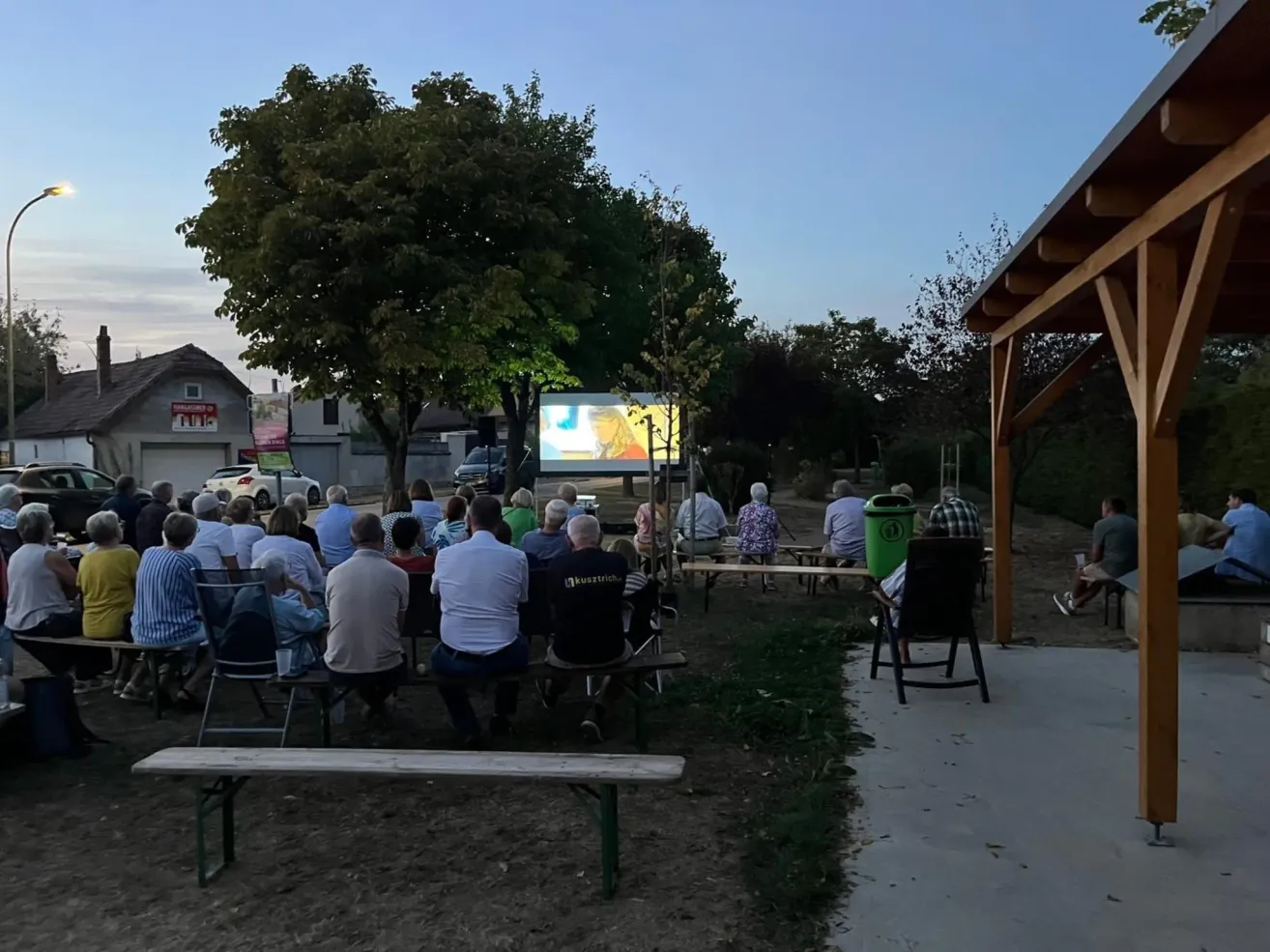 Eine Gruppe von Menschen versammelt sich im Freien, um einen auf eine Leinwand projizierten Film zu sehen. Sie sitzen auf Bänken und Stühlen, mit Bäumen und einem Gebäude im Hintergrund.