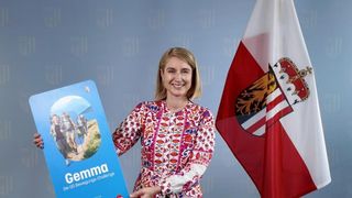A woman holds a book titled 'Gemma' with a flag behind her. The book cover features a group of people hiking.
