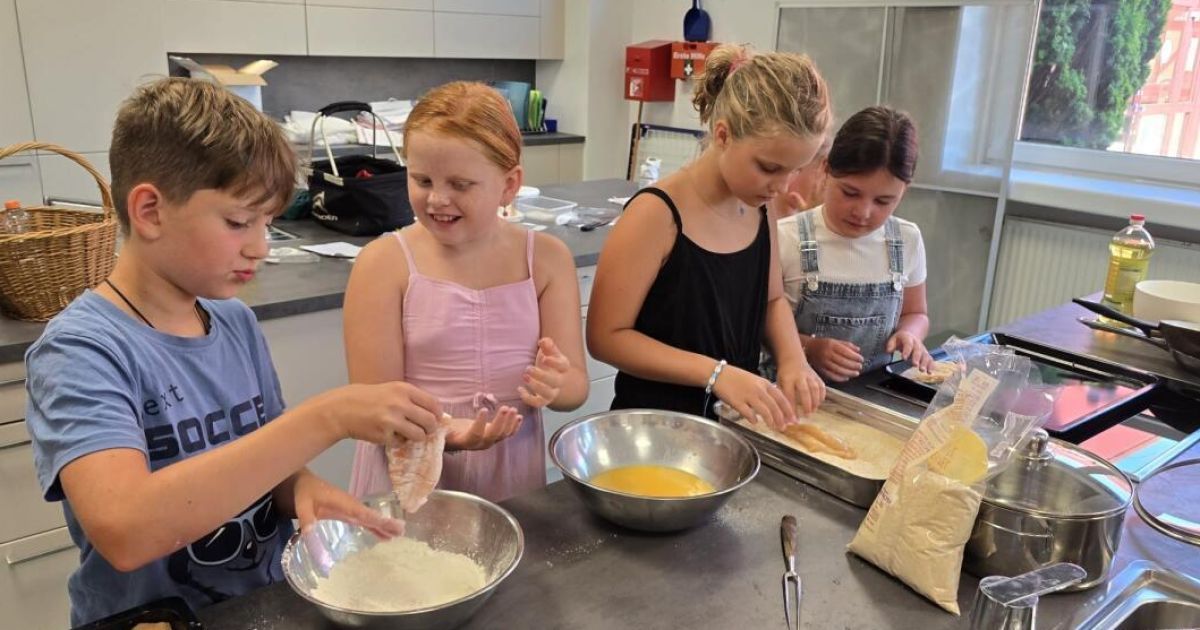Vier Kinder in einer Küche, zwei Mädchen panieren Hühnchen, während die anderen beiden Zutaten in Schüsseln mischen.