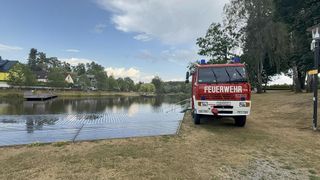 Ein roter Feuerwehrwagen steht in der Nähe eines Sees mit einer Metallrampe, die zum Wasser führt. Der Wagen hat das Wort 'Feuerwehr' auf der Vorderseite.