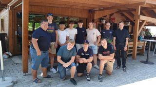 Eine Gruppe junger Männer und älterer Männer posiert für ein Foto vor einem Holzgebäude. Sie alle lächeln.