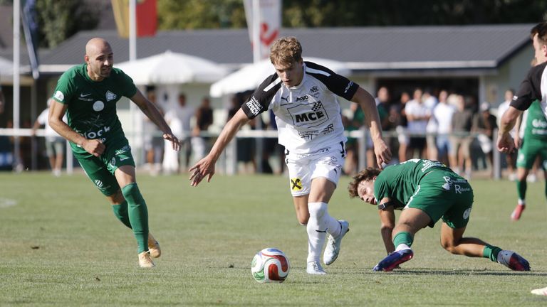 Ein Fußballspieler in einem weißen Trikot jagt einem Ball hinterher, während ein anderer Spieler in Grün zu Boden fällt.