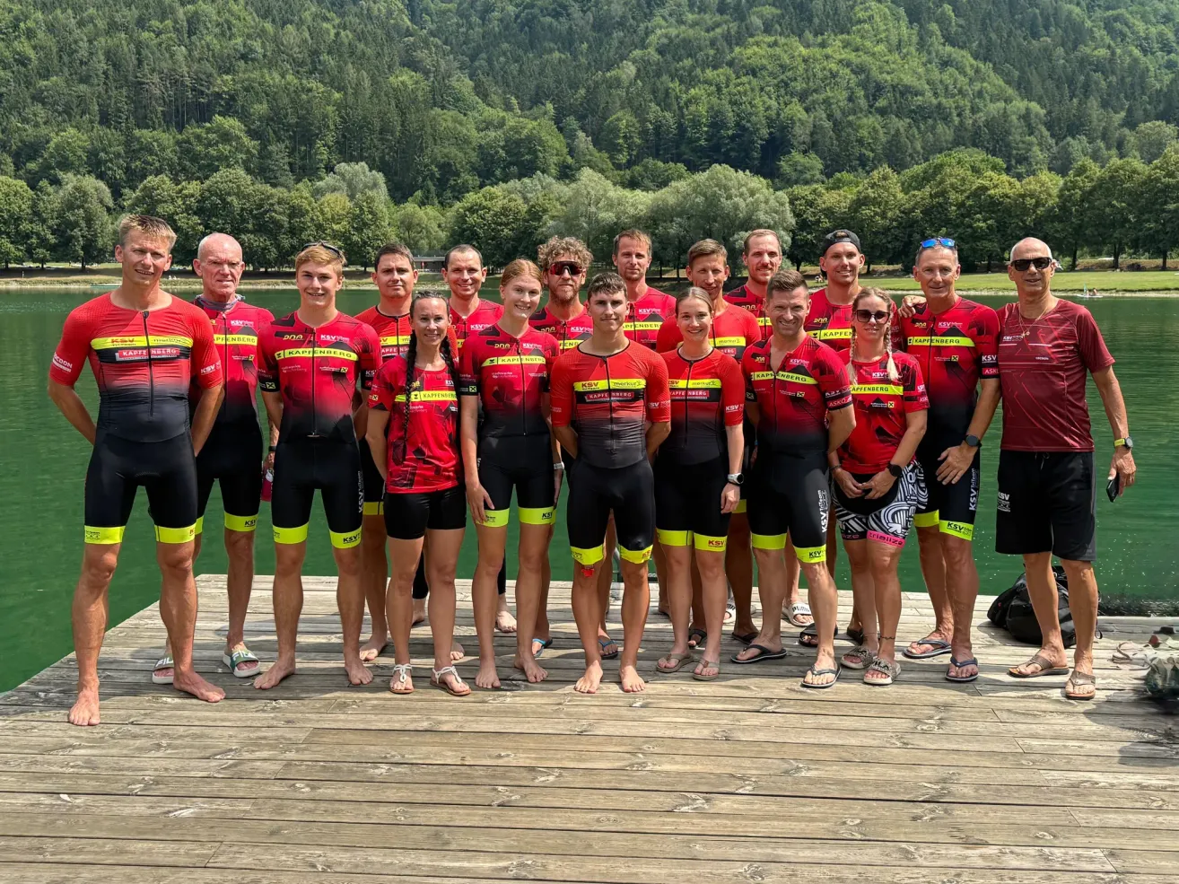 Eine Gruppe von Menschen in roten und schwarzen Radsporttrikots steht auf einem Steg am Wasser, mit Bergen im Hintergrund.