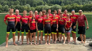Eine Gruppe von Menschen in roten und schwarzen Radsporttrikots steht auf einem Steg am Wasser, mit Bergen im Hintergrund.