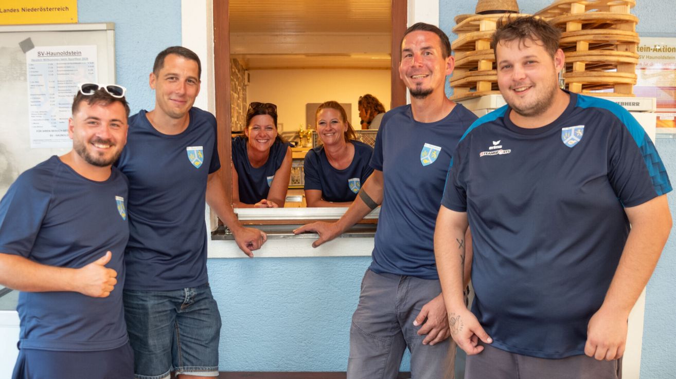 Vier Personen in gleichen blauen T-Shirts stehen vor einem Fenster und lächeln. Zwei Frauen sind im Fenster, eine mit Sonnenbrille auf dem Kopf.