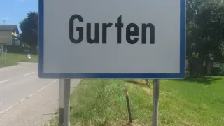 A white sign with blue borders reads 'Gurten' on a grassy roadside. Behind it, a house and trees are visible.