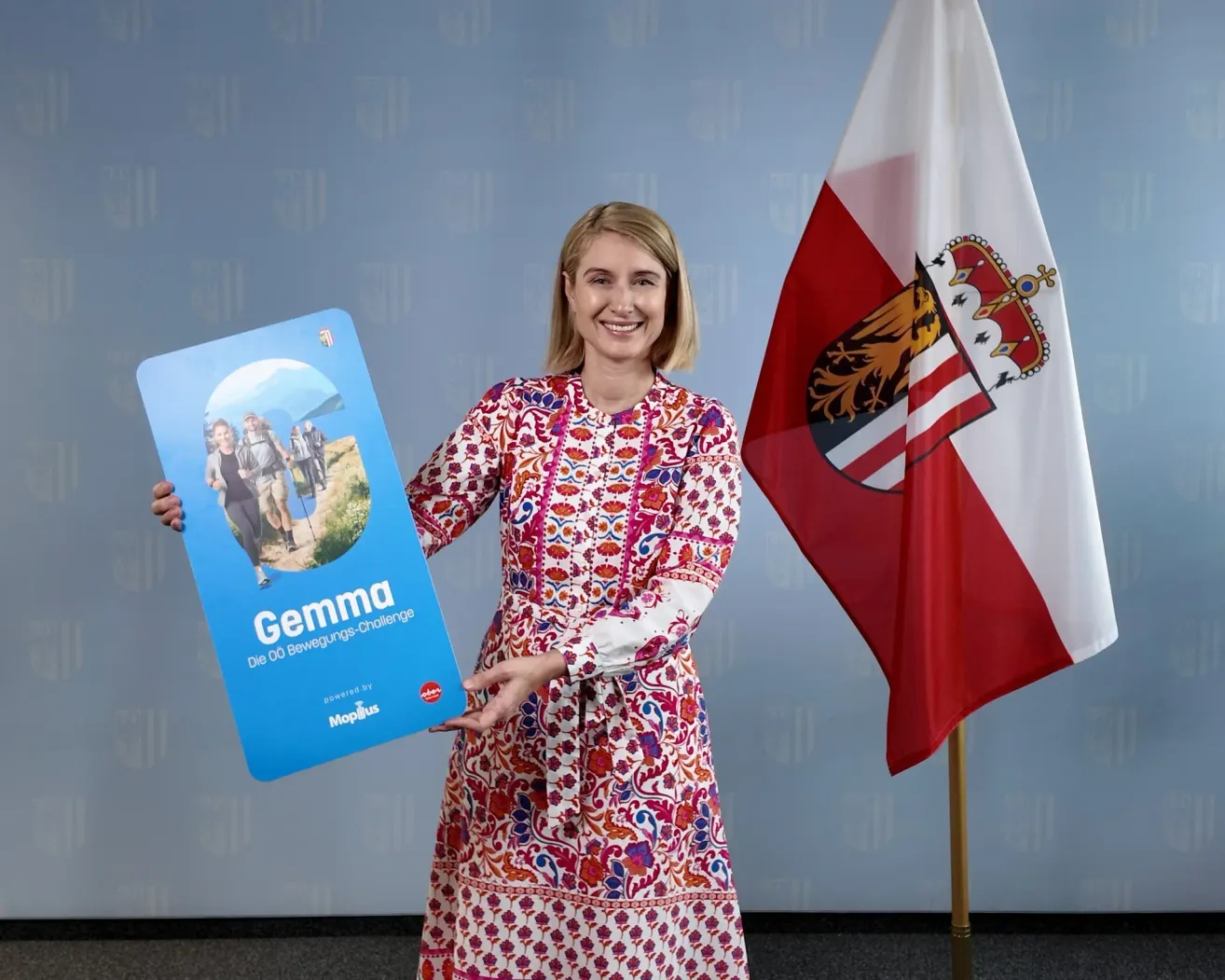 A woman in a floral dress holds a blue sign with the word Gemma and a picture of people walking. She is standing next to a flag with a coat of arms.