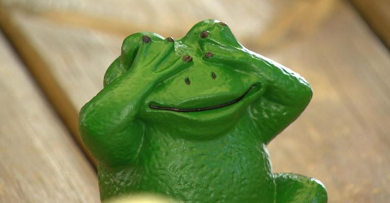 A green frog figurine with a wide smile, covering its eyes with its hands. The figurine has a smooth, glossy finish and is placed on a wooden surface.