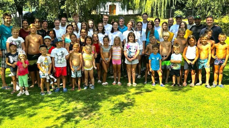 Eine große Gruppe von Kindern und Erwachsenen steht auf einem Rasenfeld und posiert für ein Gruppenfoto. Einige tragen Badekleidung, während andere lässige Outfits tragen.