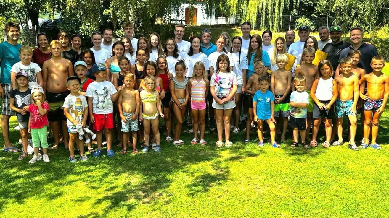 Eine große Gruppe von Kindern und Erwachsenen steht auf einem Rasenfeld und posiert für ein Gruppenfoto. Einige tragen Badekleidung, während andere lässige Outfits tragen.