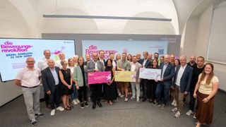 Eine Gruppe von Menschen posiert für ein Foto in einem Raum mit einem Banner, das 'die Bewegungs-Akademie' sagt. Sie halten Zertifikate und lächeln.