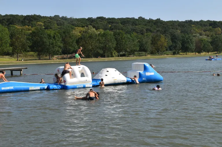 A group of people are enjoying a water obstacle course on a lake with trees in the background.