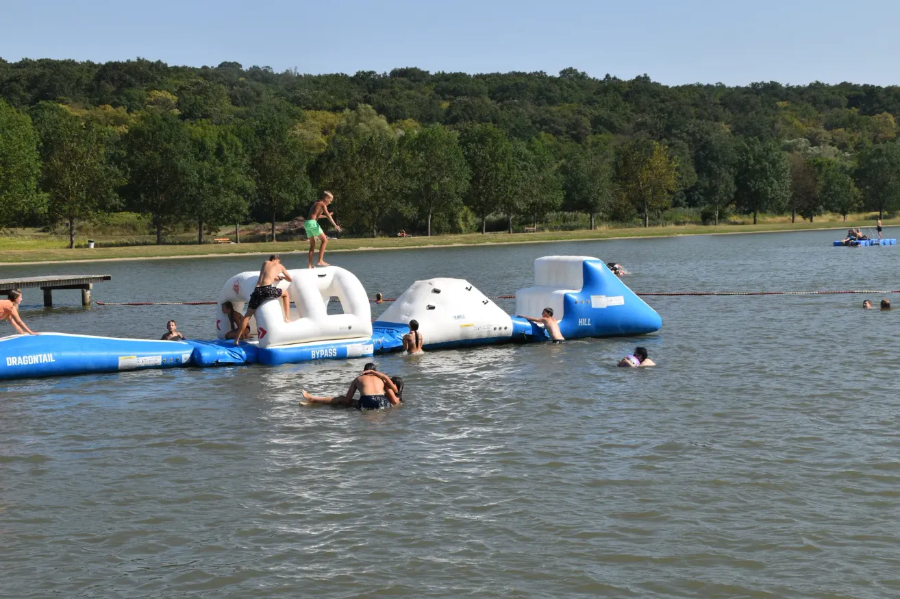 A group of people are enjoying a water obstacle course on a lake with trees in the background.