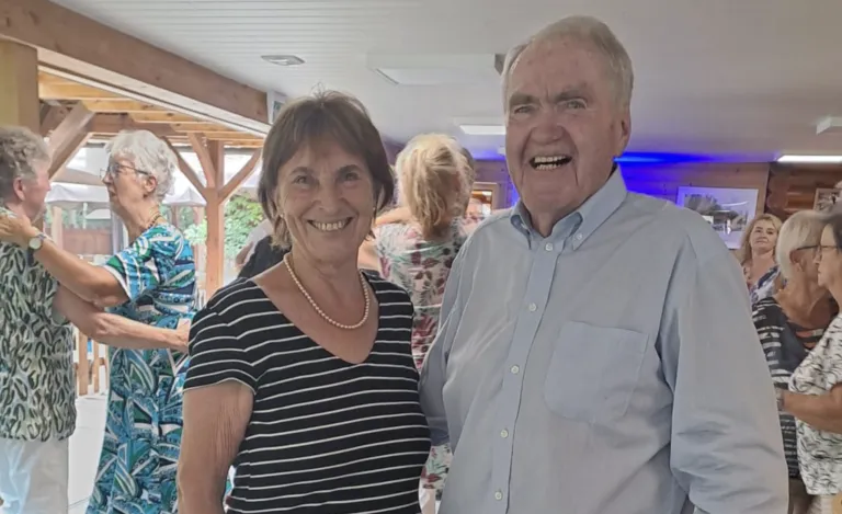 An elderly man and woman are smiling and posing for a photo at an indoor event. The man is wearing a light blue shirt, and the woman is wearing a black and white striped shirt. Behind them, a few people are standing, and the background is blurred.