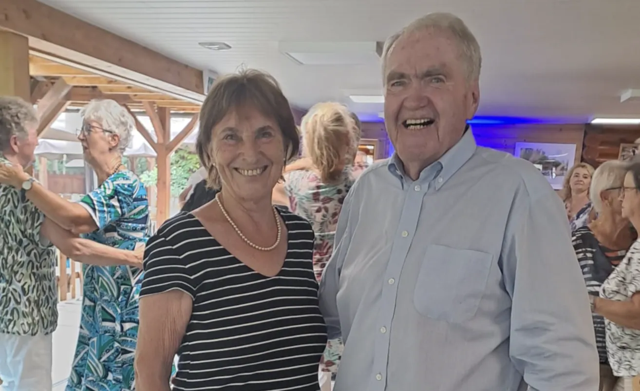 An elderly man and woman are smiling and posing for a photo at an indoor event. The man is wearing a light blue shirt, and the woman is wearing a black and white striped shirt. Behind them, a few people are standing, and the background is blurred.