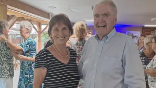 An elderly man and woman are smiling and posing for a photo at an indoor event. The man is wearing a light blue shirt, and the woman is wearing a black and white striped shirt. Behind them, a few people are standing, and the background is blurred.