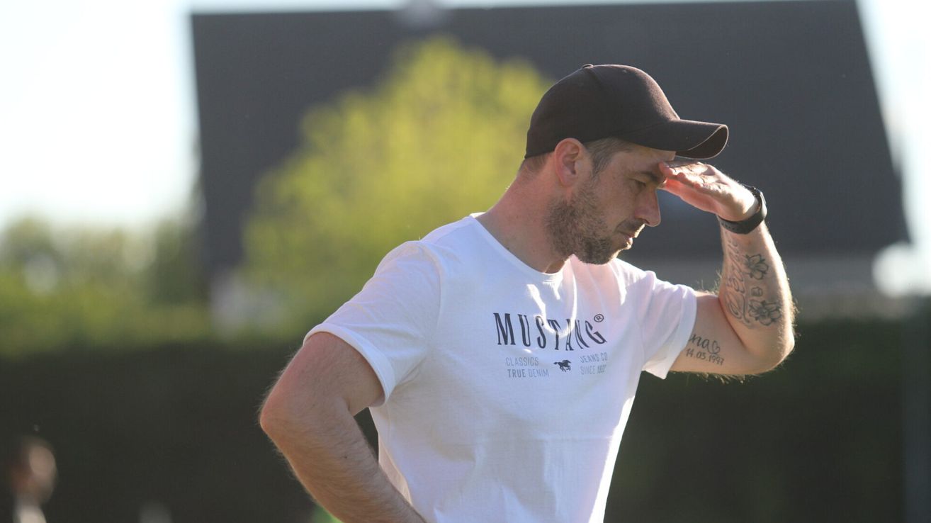 Ein Mann in einem weißen T-Shirt mit dem Wort 'Mustang' darauf, trägt eine schwarze Kappe und eine Uhr und steht draußen.