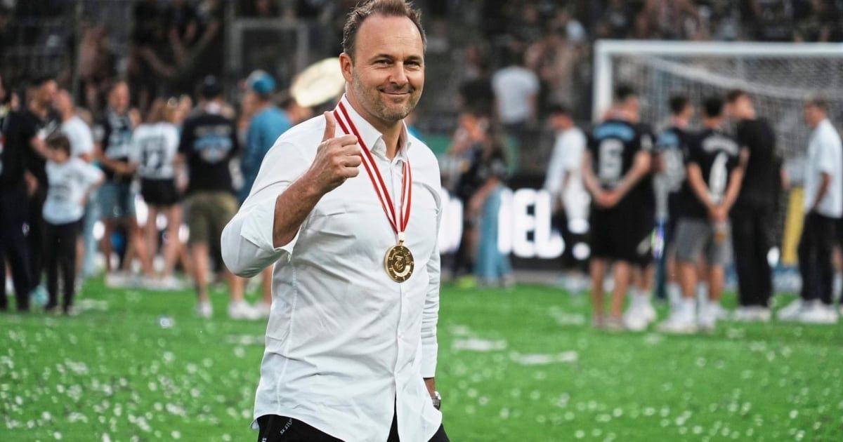 Ein Mann in einem weißen Hemd mit einer goldenen Medaille um den Hals gibt auf einem Fußballfeld einen Daumen hoch.