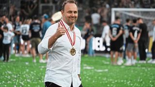 Ein Mann in einem weißen Hemd mit einer goldenen Medaille um den Hals gibt auf einem Fußballfeld einen Daumen hoch.