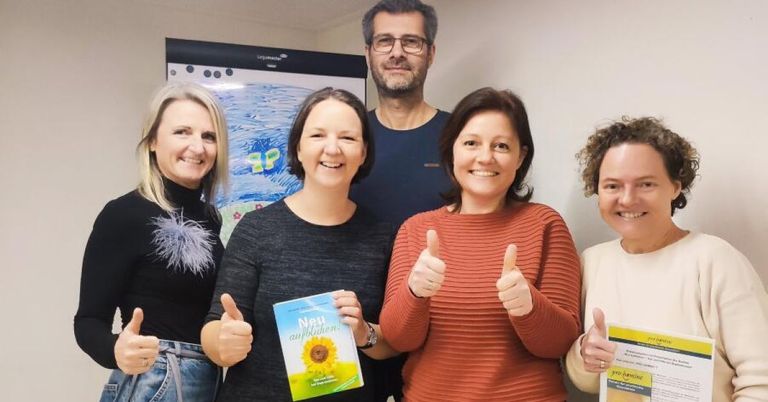 A group of five people are smiling and giving thumbs up. A woman in the middle is holding a book titled 'Neue Aufplodung'. Behind them is a whiteboard with a drawing of a butterfly.