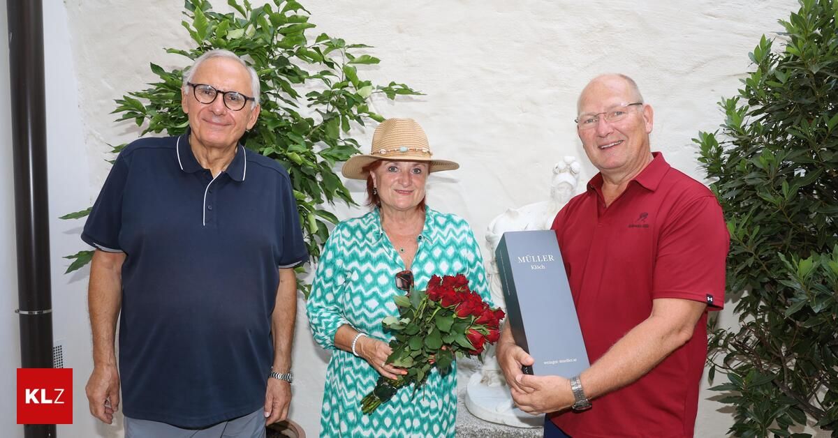 Drei Menschen stehen lächelnd nebeneinander, einer hält einen Strauß roter Rosen und ein anderer eine blaue Box mit der Aufschrift Muller. Sie stehen vor einer weißen Wand mit einer Pflanze und einer Statue.