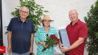 Drei Menschen stehen lächelnd nebeneinander, einer hält einen Strauß roter Rosen und ein anderer eine blaue Box mit der Aufschrift Muller. Sie stehen vor einer weißen Wand mit einer Pflanze und einer Statue.