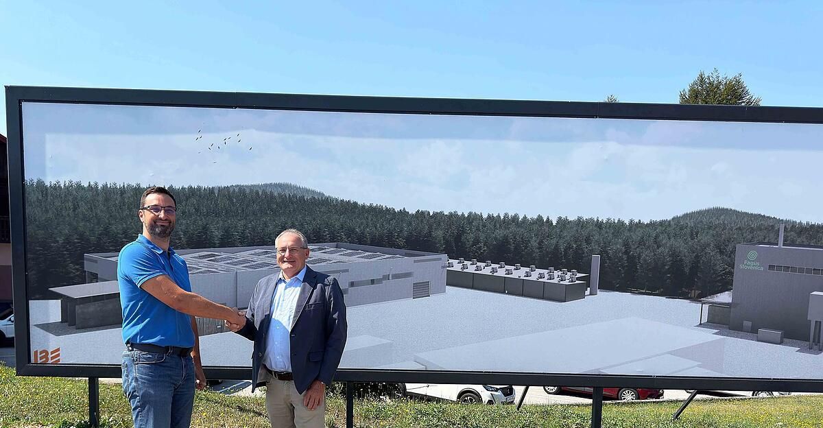 Two men stand in front of a large billboard showing a factory with solar panels on the roof. The man on the left wears a blue shirt and jeans, while the man on the right wears a navy blazer and light pants.