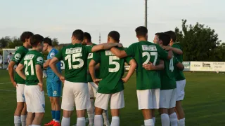A soccer team in green jerseys with numbers 5, 15, and 27 huddles together on the field under a clear sky.