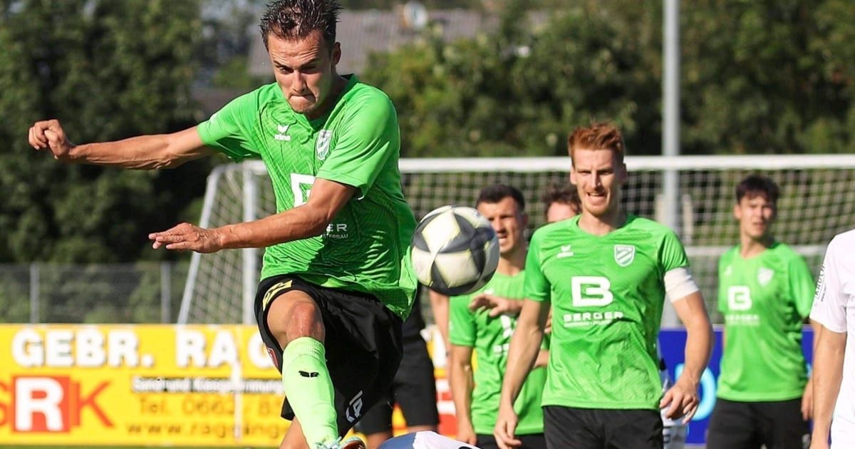 Ein Fußballspieler in einem grünen Trikot tritt einen Ball in der Luft während eines Spiels. Dahinter beobachten zwei Teamkollegen. Im Hintergrund befindet sich ein Tornetz und ein Banner mit Text.