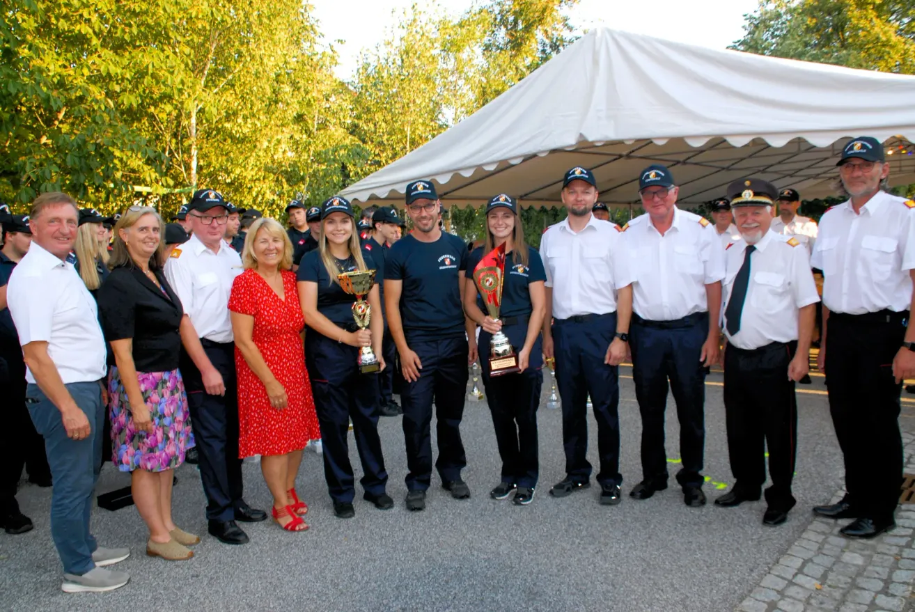 Eine Gruppe von Menschen, darunter Männer und Frauen, steht unter einem weißen Zelt, hält Trophäen und posiert für ein Foto. Sie sind alle in Uniform gekleidet.