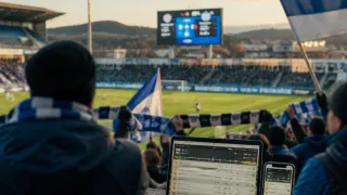 Ein Fußballfan beobachtet ein Spiel auf einer großen Leinwand, während er eine Fahne hält. Die Anzeigetafel ist auf dem Bildschirm sichtbar, und das Spielfeld ist mit Zuschauern gefüllt.