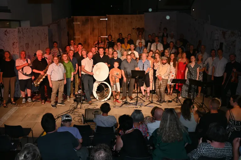 A large group of musicians and audience members are gathered on a stage, with musical instruments and equipment visible. The musicians are standing and playing various instruments, while the audience is seated in front of them.