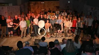 Eine große Gruppe von Musikern und Zuschauern versammelt sich auf einer Bühne, mit sichtbaren Musikinstrumenten und Ausrüstung. Die Musiker stehen und spielen verschiedene Instrumente, während das Publikum vor ihnen sitzt.