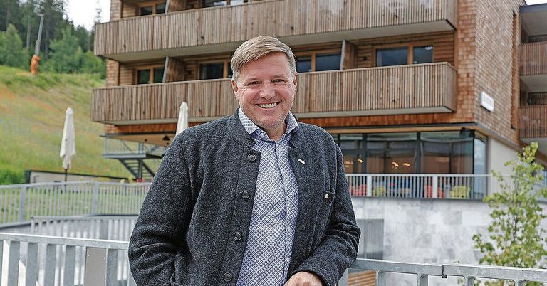 Ein Mann mit fröhlicher Miene steht vor einem hölzernen Gebäude mit Balkonen und Fenstern, gekleidet in eine graue Strickjacke und ein blaues kariertes Hemd.