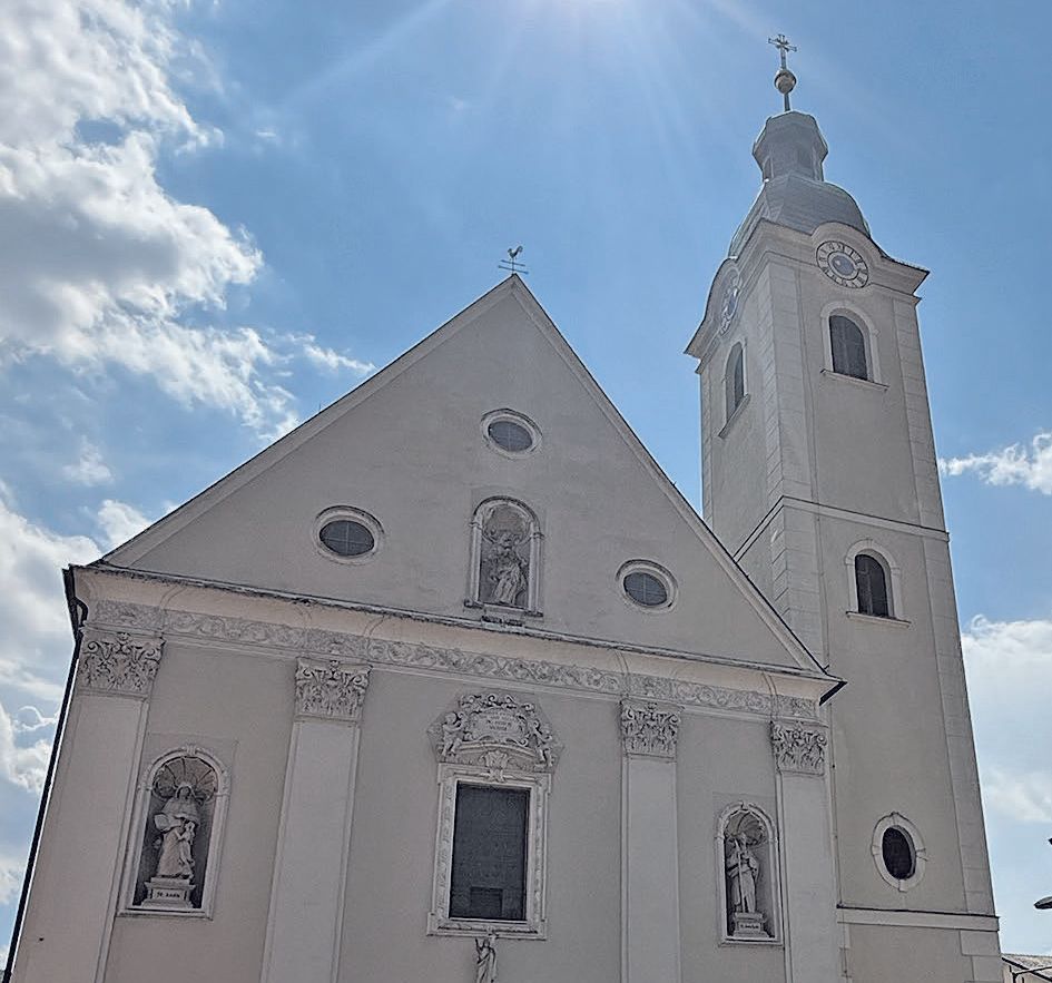 A white church with a tall tower, adorned with statues and intricate designs, under a bright sky with clouds.
