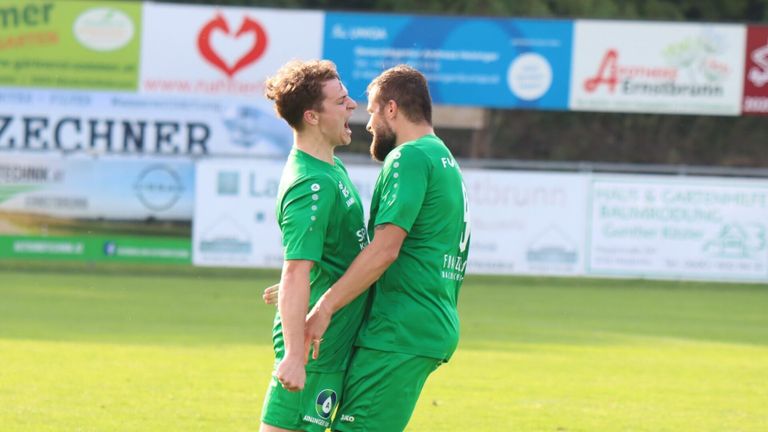 Zwei Männer in grünen Fußballtrikots feiern auf einem Feld mit einem verschwommenen Hintergrund von Werbung.