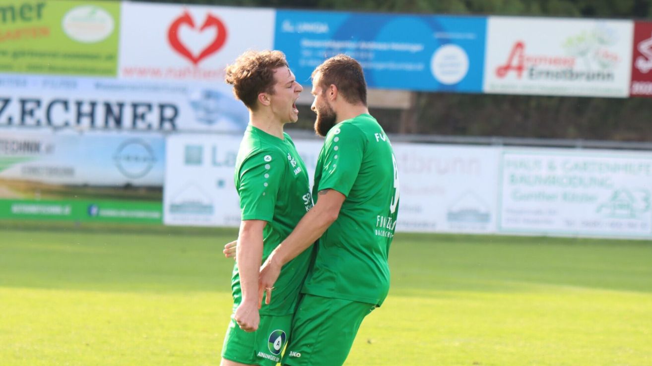 Zwei Männer in grünen Fußballtrikots feiern auf einem Feld mit einem verschwommenen Hintergrund von Werbung.