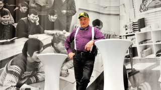 Ein älterer Mann in einem lila Hemd und Hosenträgern steht vor einer Wand mit einem Foto von Schülern in einem Klassenzimmer.