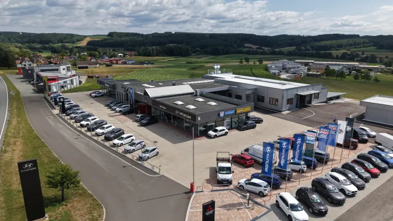 Aerial view of a car dealership with multiple cars parked in front and around the building. The dealership is surrounded by a green field and a cloudy sky.