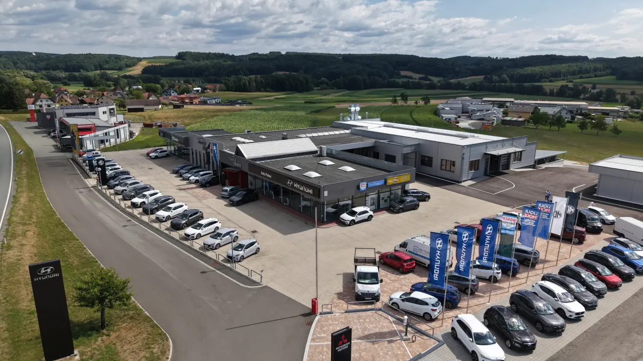 Luftaufnahme eines Autohauses mit mehreren Autos vor und um das Gebäude geparkt. Das Autohaus ist von einem grünen Feld und einem bewölkten Himmel umgeben.