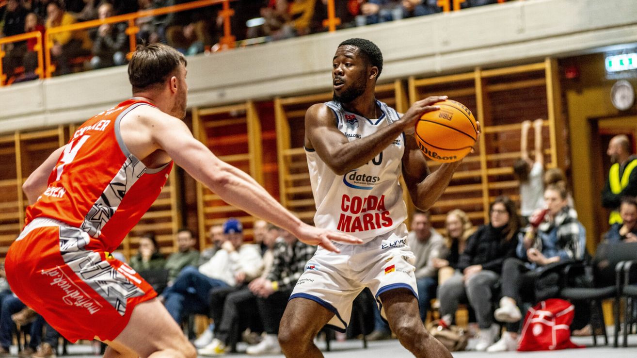 Ein Basketballspieler in einem weißen Trikot mit 'Colda Maris' darauf dribbelt den Ball, während ein anderer Spieler in Rot versucht, ihn zu blockieren. Zuschauer sehen von den Tribünen zu.