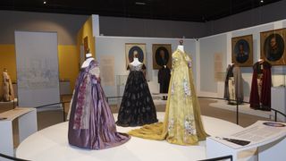 Drei Vintage-Kleider auf Mannequins in einer Museumsausstellung, mit einem gelben Kleid im Vordergrund, einem schwarzen Kleid in der Mitte und einem lila Kleid hinten. Hinter den Kleidern hängen zwei gerahmte Porträts an der Wand.