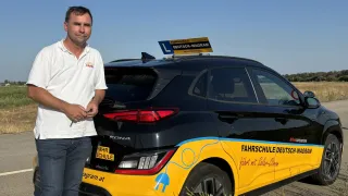 A man stands next to a black and yellow Kona electric car with a yellow sign on top that reads Fahrschule Deutsch-Wagram.