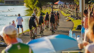 Eine Gruppe von Radfahrern mit Helmen und Radfahreranzügen fährt auf einem Weg am Fluss entlang, mit Zuschauern und einem Zelt im Hintergrund.