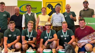 A group of people, some wearing green shirts and medals, are posing for a photo with buckets and flowers in front of them. Behind them, there are banners with logos and text.