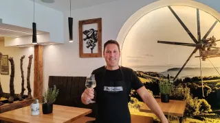 A man in a black apron stands in a restaurant, holding a glass of white wine. Behind him is a wall with a large clock and a picture frame. The room has wooden tables and chairs, and there are plants on the tables.