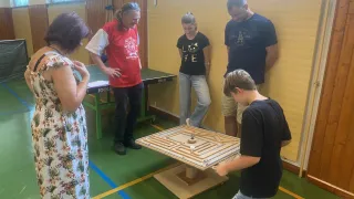 A group of people are gathered around a large wooden maze game, with a boy kneeling and playing while others watch.